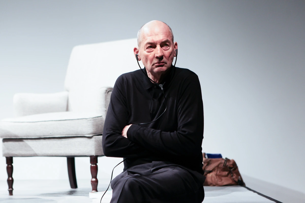 Rem Koolhaas seated on stage in a black outfit with an earpiece, photographed against a minimal light background