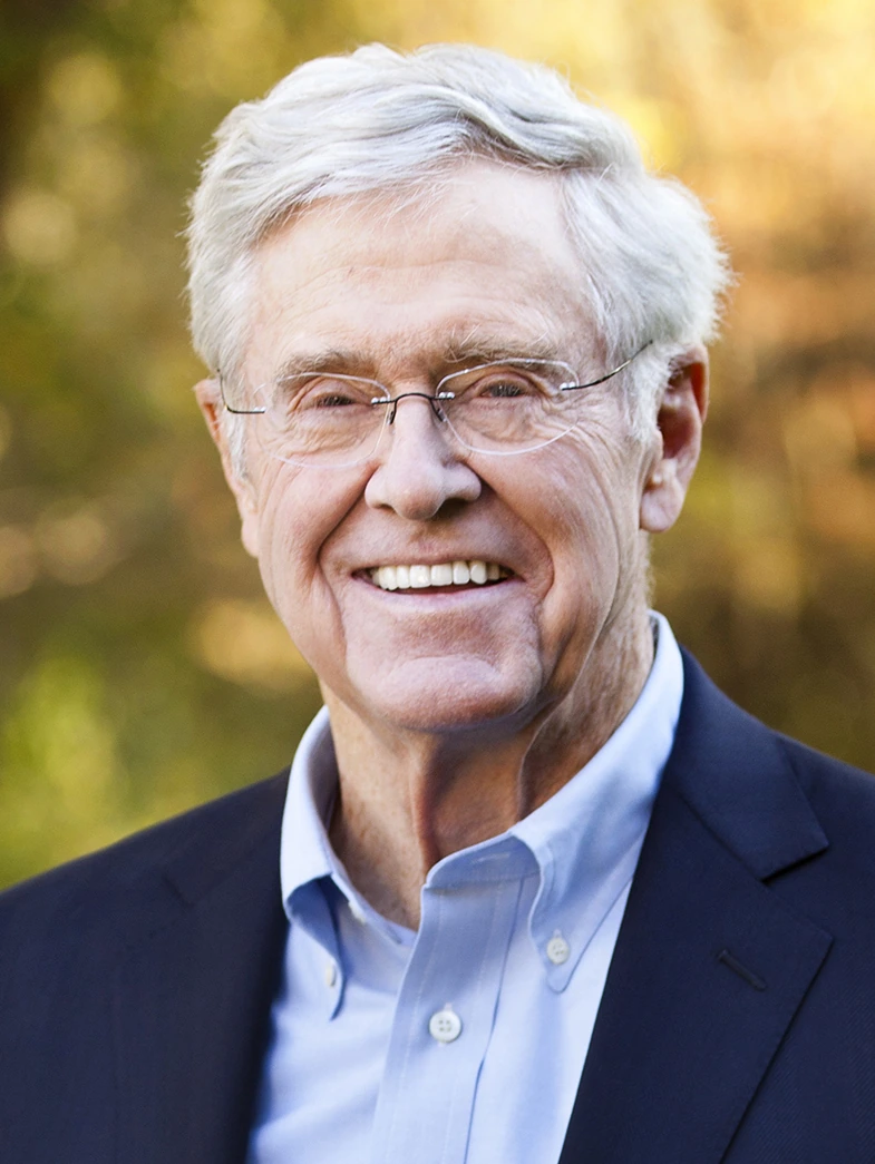 Portrait photo of Charles Koch smiling in a suit and glasses against a blurred outdoor background.